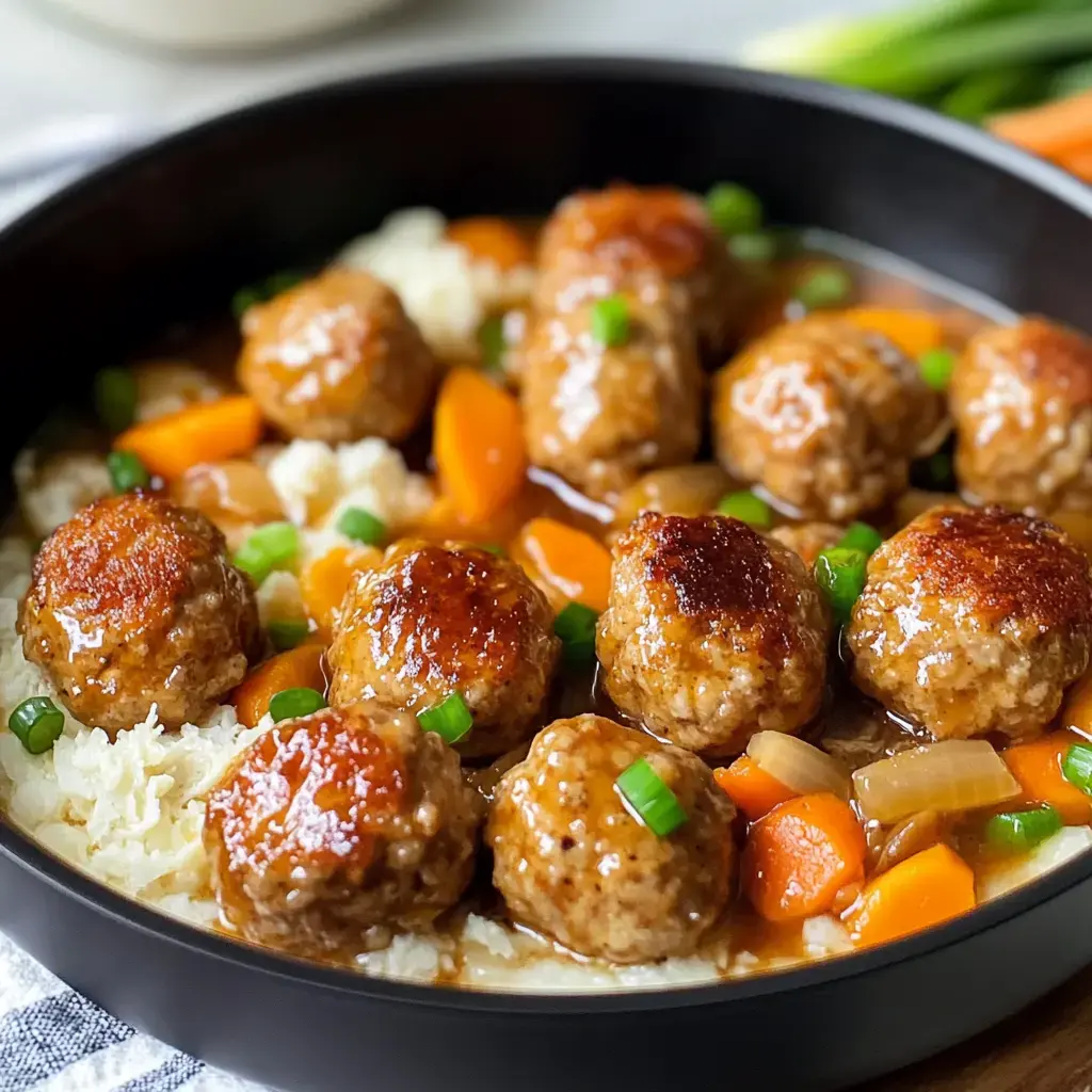 A black bowl filled with meatballs in a savory sauce, surrounded by carrots, onions, and garnished with green onions on a bed of cauliflower rice.