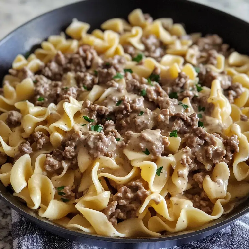 A skillet filled with egg noodles topped with ground beef in a creamy sauce, garnished with parsley.