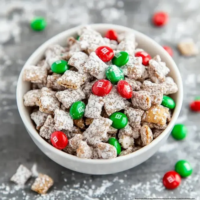 A bowl filled with a sweet snack mix of cereal pieces coated in powdered sugar, featuring red and green candy-coated chocolates.