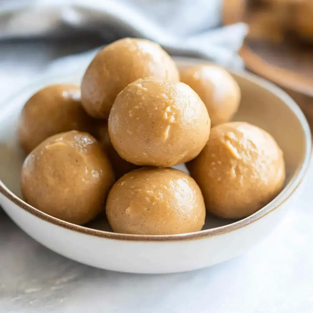 A bowl filled with several round, beige energy balls sits on a textured surface.