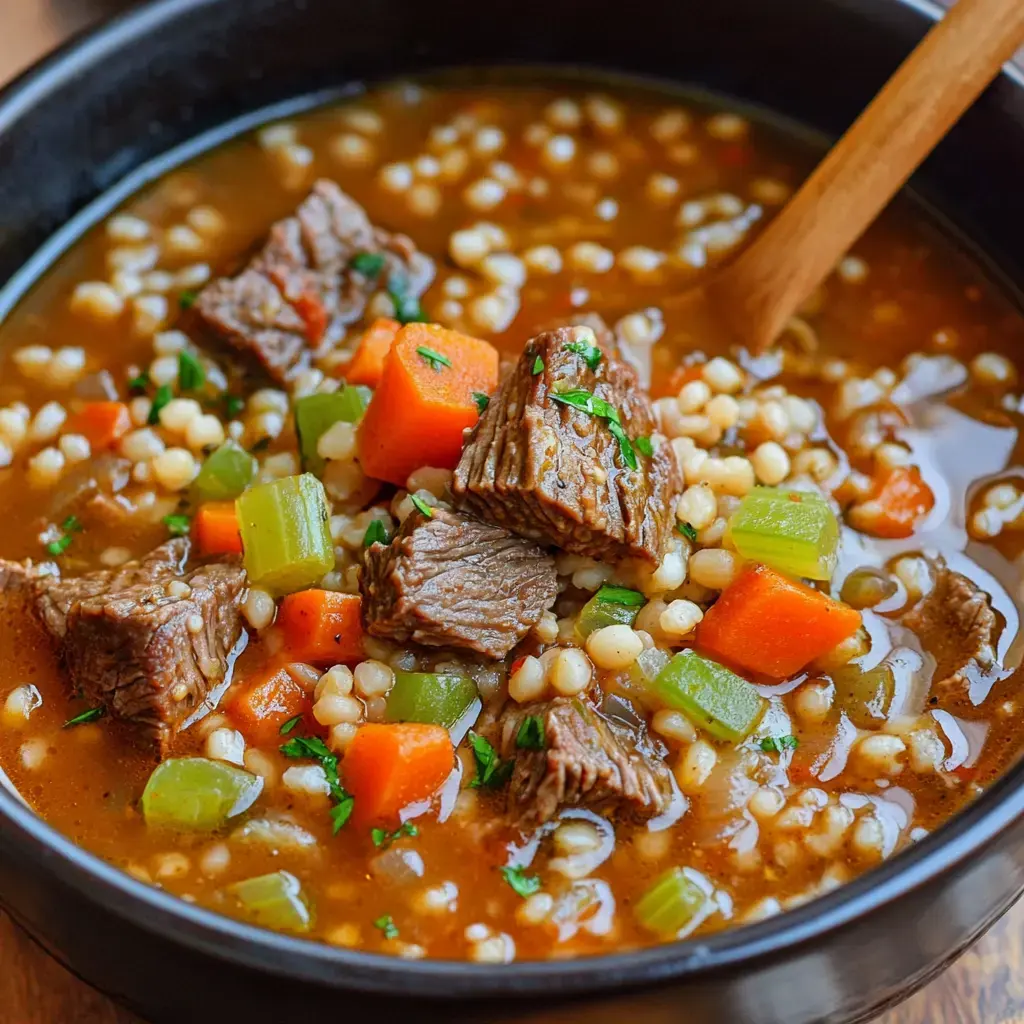 A close-up of a hearty beef soup with barley, featuring chunks of beef, carrots, celery, and garnished with parsley in a black bowl.