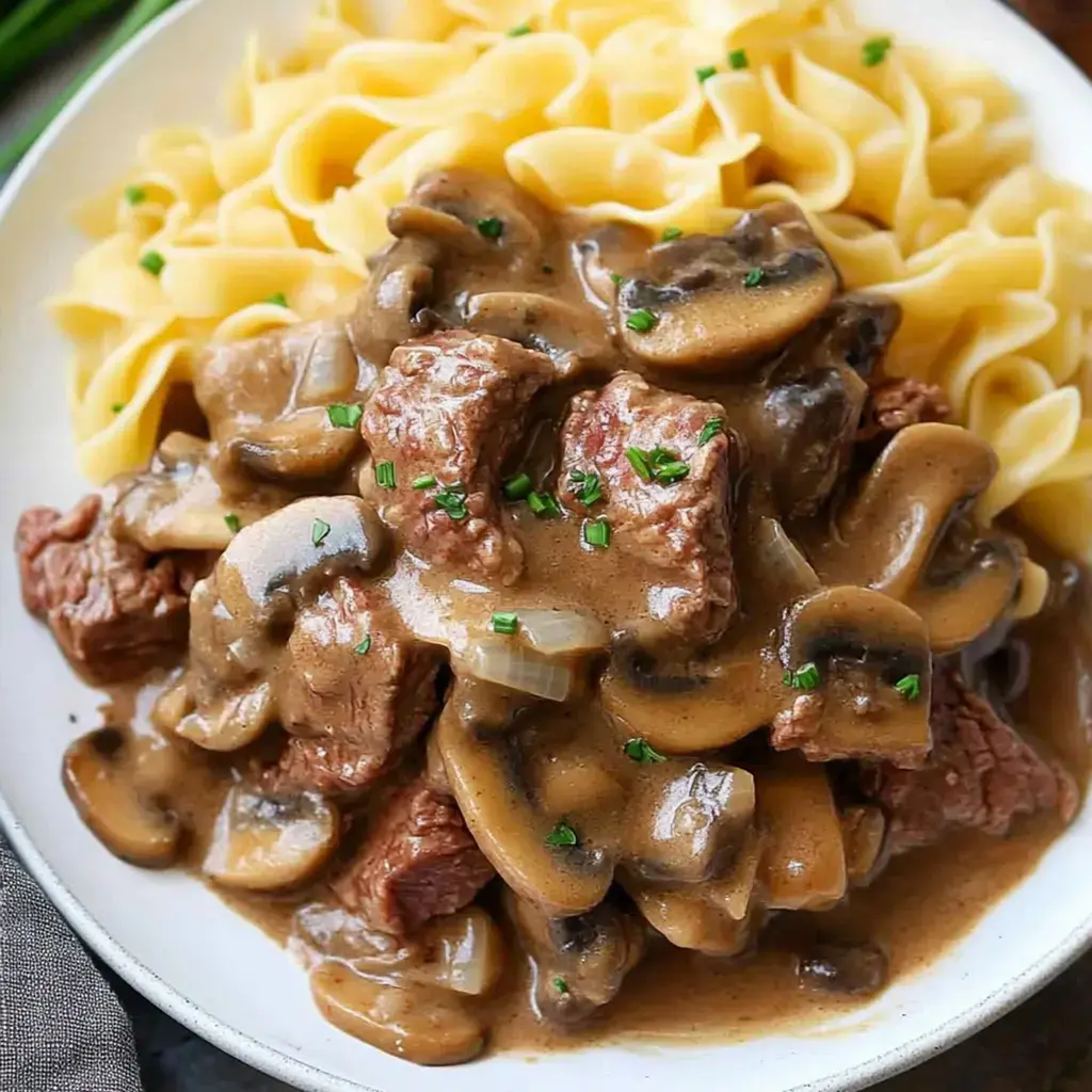 A bowl of egg noodles served with tender beef chunks in a creamy mushroom sauce, garnished with chopped green onions.