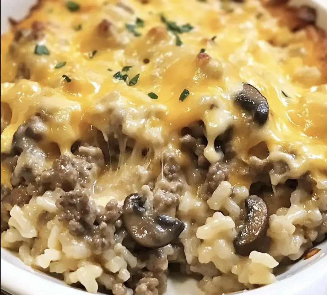 A close-up of a cheesy casserole featuring ground beef, mushrooms, and rice topped with melted cheddar cheese and garnished with parsley.