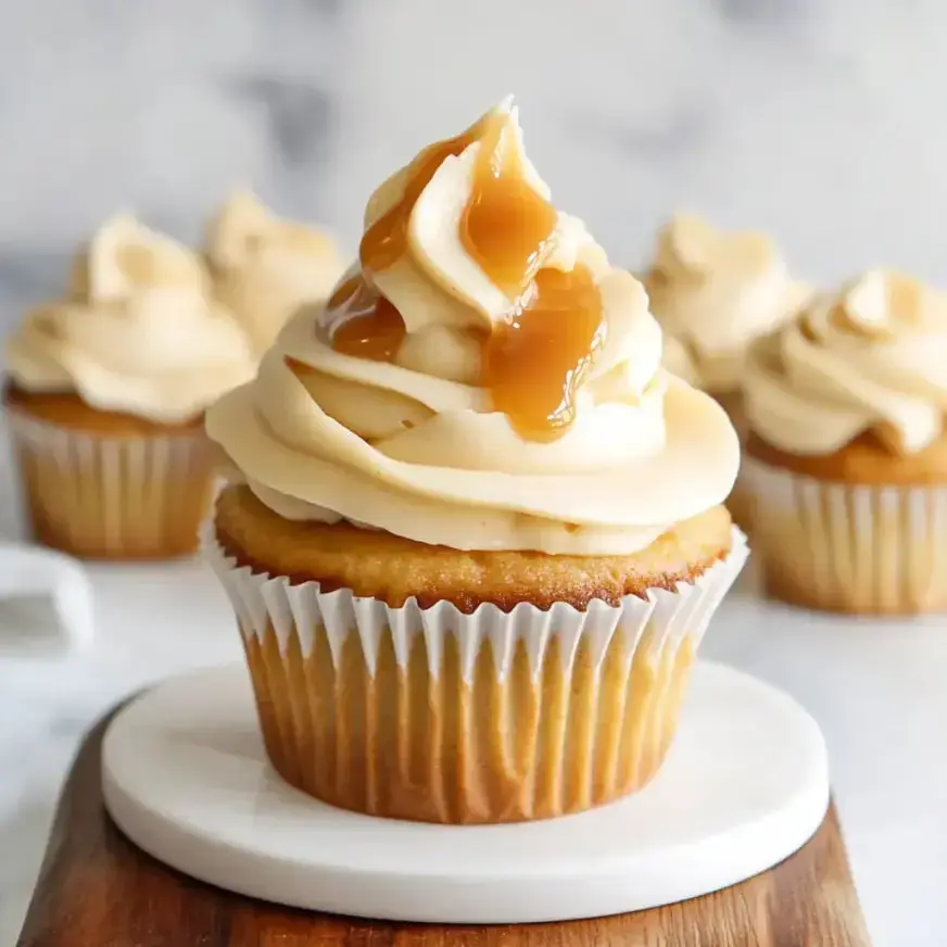 A close-up of a gourmet cupcake topped with swirls of cream frosting and a drizzle of caramel sauce, placed on a white circular plate.