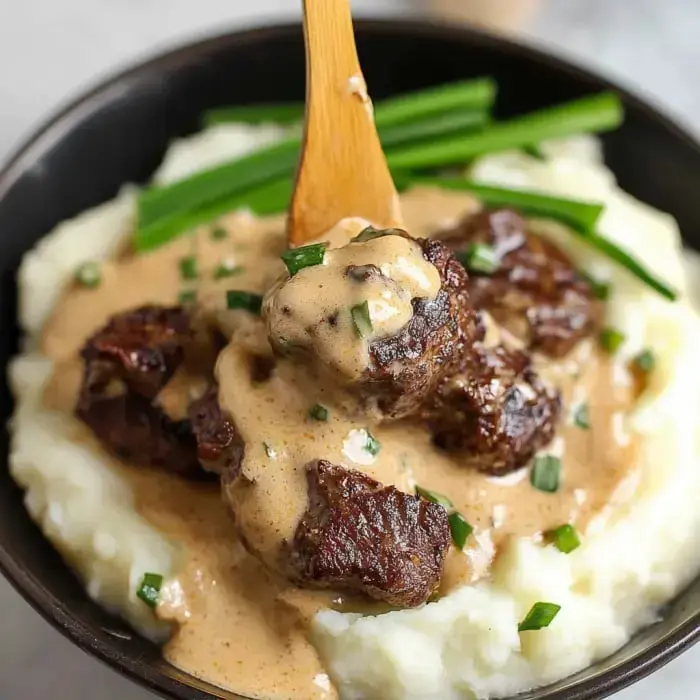 A bowl of creamy mashed potatoes topped with tender beef pieces in a savory sauce, garnished with green onions.