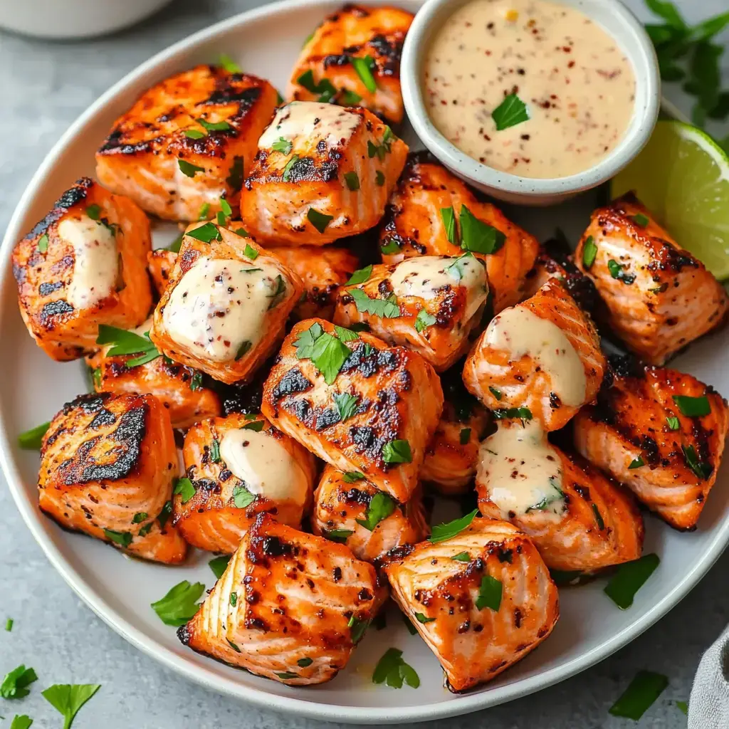 A plate of grilled salmon cubes garnished with parsley and served with a small bowl of sauce and a lime wedge.