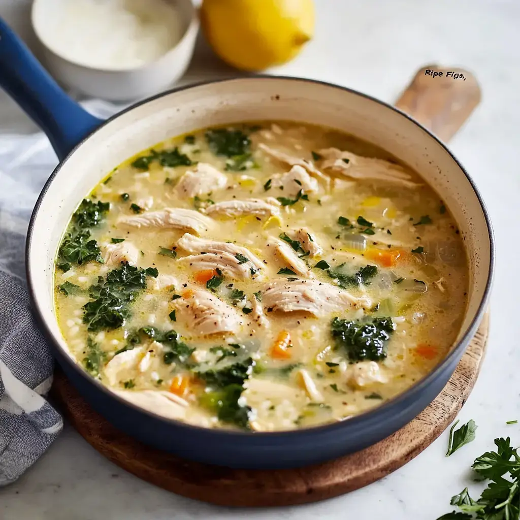 A bowl of chicken soup with shredded chicken, kale, and colorful vegetables on a wooden surface, accompanied by a lemon and a small bowl of sauce.