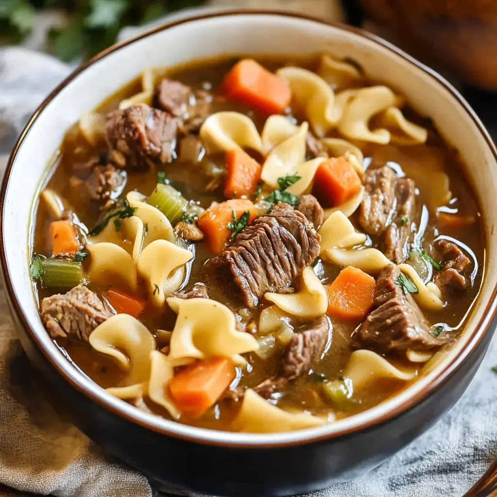 A bowl of hearty beef noodle soup featuring tender chunks of beef, carrots, celery, and egg noodles in a savory broth.