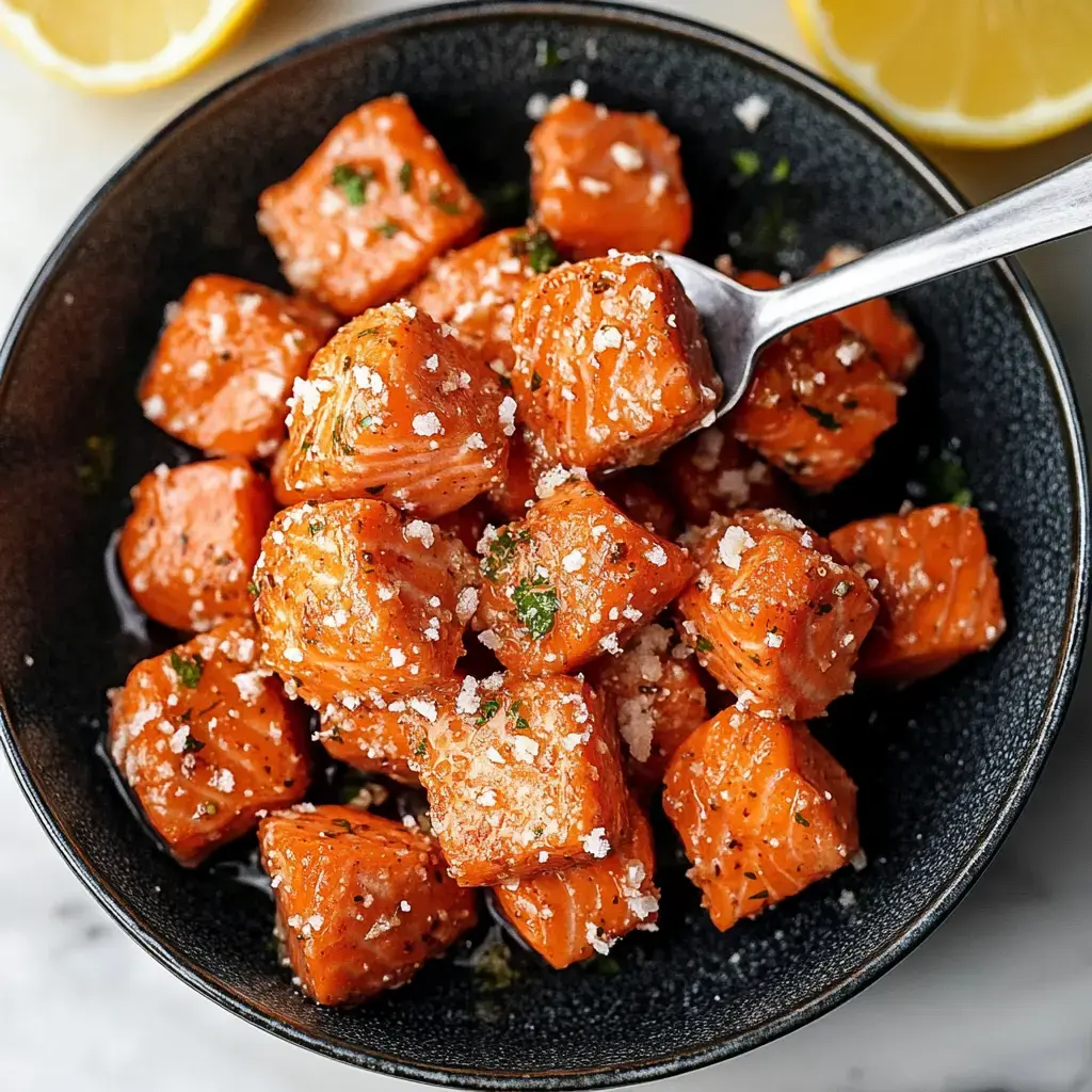 A close-up of a black bowl filled with diced, seasoned salmon garnished with herbs and salt, accompanied by lemon slices.