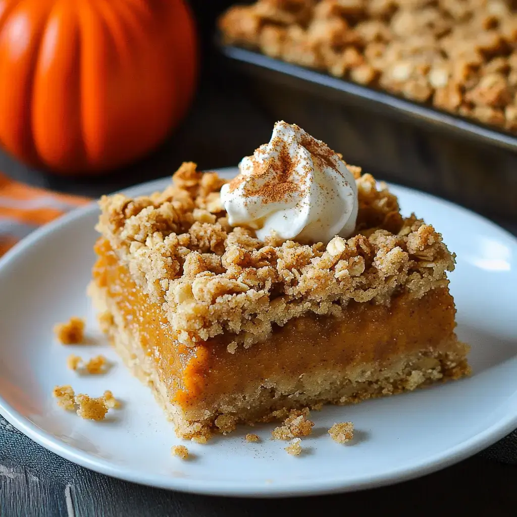 A slice of pumpkin dessert topped with whipped cream and cinnamon, served on a white plate, with a small pumpkin in the background.