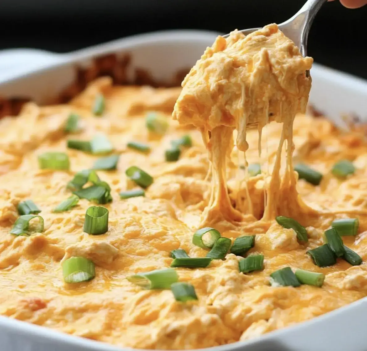 A serving spoon lifts a cheesy, creamy dish topped with green onions from a casserole dish.