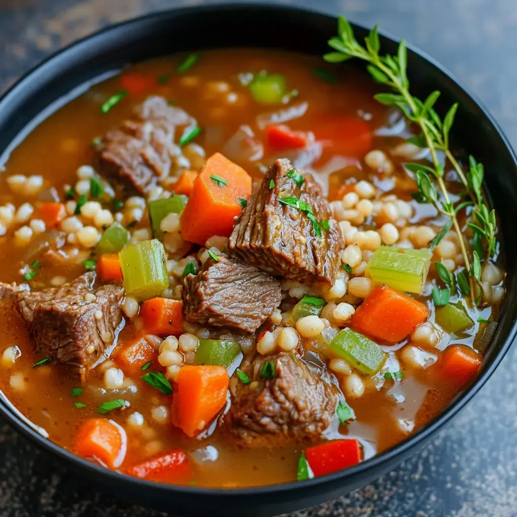 A close-up view of a bowl of hearty beef stew containing chunks of meat, carrots, celery, and pearl couscous, garnished with fresh herbs.