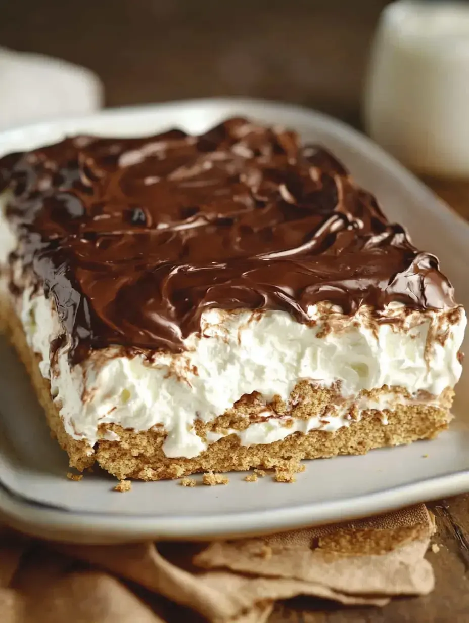 A slice of layered dessert featuring a graham cracker base topped with whipped cream and chocolate frosting, served on a white plate.