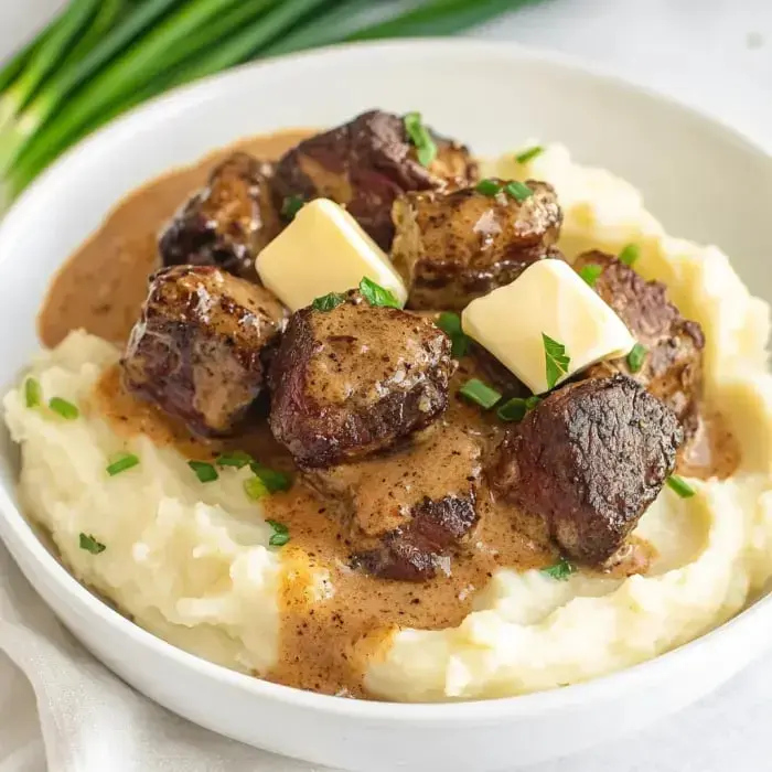 A bowl of creamy mashed potatoes topped with chunks of beef in gravy and pats of butter, garnished with chopped green onions.