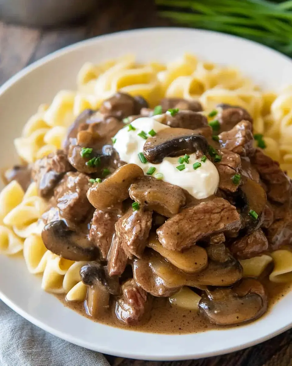 A plate of egg noodles topped with beef, mushrooms, and a dollop of sour cream, garnished with chopped chives.