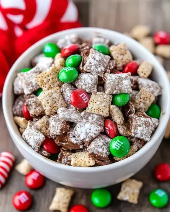 A bowl of holiday snack mix containing chocolate-coated cereal, powdered sugar, and red and green candy-coated chocolates.