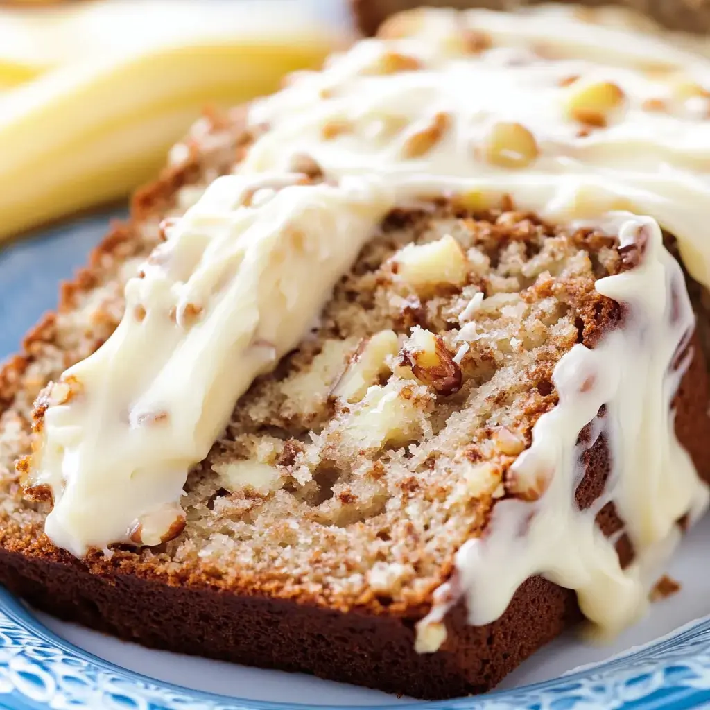 A slice of banana bread topped with creamy frosting and sprinkled with nuts, served on a blue patterned plate alongside bananas.