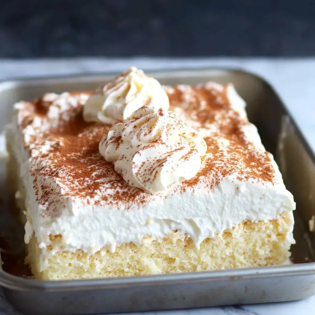 A rectangular cake topped with whipped cream and a dusting of cocoa powder sits in a metal baking dish.