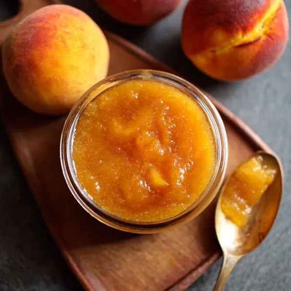 A glass of peach jam on a wooden tray.