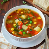 A bowl of colorful vegetable soup with various ingredients including corn, peas, potatoes, and carrots, garnished with herbs.