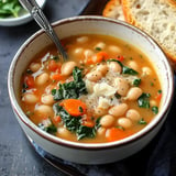 A bowl of hearty white bean soup with carrots, kale, and a sprinkle of cheese, accompanied by slices of bread.