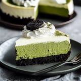 A slice of matcha cheesecake with layers of green tea filling and whipped cream, topped with an Oreo cookie, served on a dark plate.