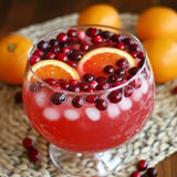 A glass bowl filled with a vibrant red drink garnished with cranberries and orange slices, resting on a woven placemat, with whole oranges in the background.