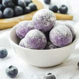 A bowl filled with purple, powdered spherical treats sits on a white surface, surrounded by scattered blueberries.