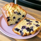 A slice of blueberry loaf cake is placed on a pink plate, showcasing its moist texture and blueberries throughout.