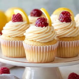 A plate of cupcakes with swirled frosting, topped with raspberries and lemon slices.