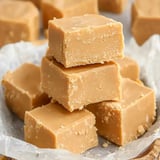 A close-up view of stacked squares of creamy caramel fudge on a textured surface.