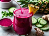 A vibrant, pink smoothie is served in a glass, garnished with a pineapple slice, alongside fresh cucumber slices, ginger, and a small bowl of beet powder.