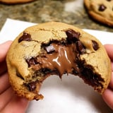 A close-up of a hand holding a chocolate chip cookie with a gooey melted chocolate filling exposed from a bite taken out.