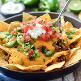 A close-up of a bowl of nachos topped with melted cheese, ground beef, jalapeños, diced tomatoes, guacamole, cilantro, and sour cream.