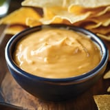 A small bowl of creamy orange dip is surrounded by tortilla chips on a wooden surface.