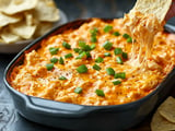 A serving of gooey, cheesy dip is being lifted with a tortilla chip, surrounded by green onions, with a bowl of chips in the background.