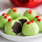 A plate of green-coated cake balls with red heart sprinkles, one partially cut in half to reveal a dark chocolate filling.