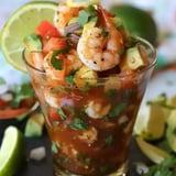 A glass filled with shrimp ceviche, featuring shrimp, diced tomatoes, avocado, and cilantro, garnished with lime and lemon slices.