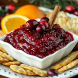 A white bowl filled with cranberry sauce garnished with cranberries and a cinnamon stick, surrounded by crackers and an orange slice.