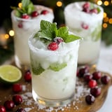 Three festive cocktails adorned with mint leaves and cranberries sit on a wooden platter, surrounded by fresh cranberries and a lime half.