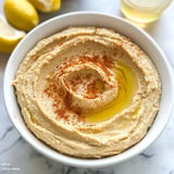 A bowl of hummus drizzled with olive oil and sprinkled with paprika, surrounded by lemon halves.