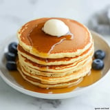 A stack of pancakes topped with a dollop of butter and drizzled with syrup, accompanied by blueberries on a plate.