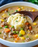 A creamy bowl of savory soup with ground beef, peas, carrots, corn, and topped with a scoop of mashed potatoes.