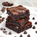 Three stacked chocolate brownies are placed on a white plate, surrounded by scattered chocolate chips.