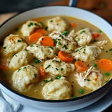 A bowl of chicken and dumplings soup features fluffy dumplings, tender chicken, and sliced carrots in a savory broth, garnished with chopped parsley.