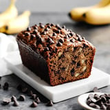 A sliced loaf of chocolate chip banana bread is displayed on a white plate, with scattered chocolate chips and ripe bananas in the background.