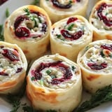 A close-up view of rolled appetizers featuring cream cheese, dried cranberries, and green onions, arranged on a white plate with decorative green herbs.