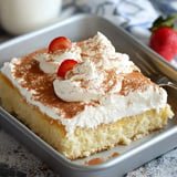 A rectangular vanilla cake topped with whipped cream, cocoa powder, and sliced strawberries on a gray baking tray.