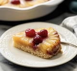 A slice of pineapple upside-down cake topped with cherries on a decorative white plate.