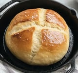 A golden-brown round loaf of bread with a crispy crust rests in a cast iron skillet.